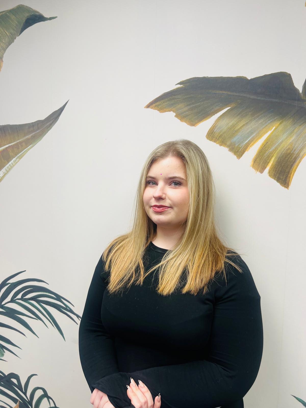 Cerys, with long blonde hair, just past her shoulders, wearing a black top in front of a botanical wallpaper
